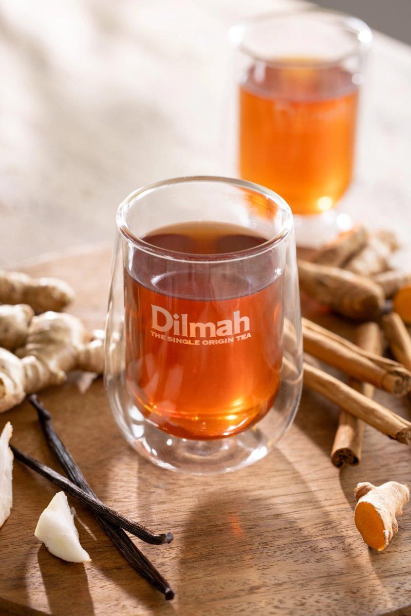 Glass of Dilmah tea surrounded by ginger, cinnamon, vanilla and turmeric